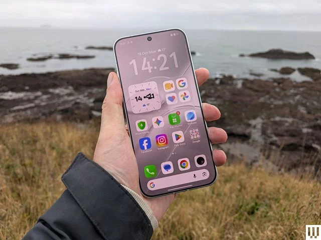 Hand holding a slim mobile phone with the screen showing apps a rocky coastline in the background