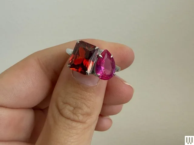 Person's hand pinching a ring with two gemstones, pink and red